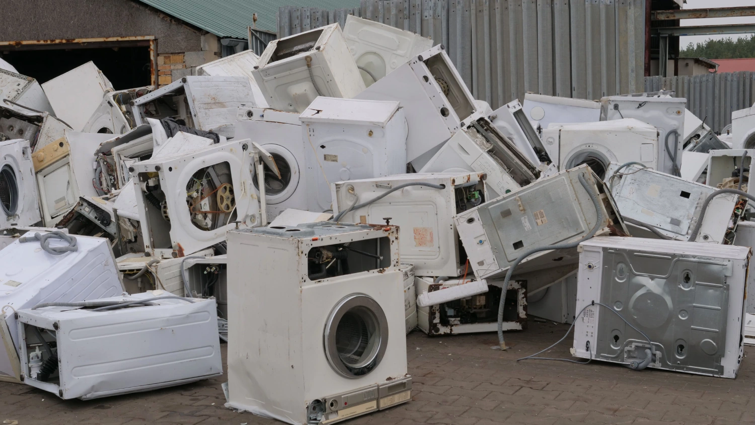 A large pile of scrapped white washing machines and dryers stacked at an outdoor appliance recycling center.